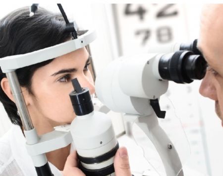 A imagem mostra uma mulher realizando um exame de vista em um consultório oftalmológico. Ela está sentada em frente a um equipamento usado para avaliar os olhos, chamado lâmpada de fenda. Um profissional da saúde, parcialmente visível, observa através do aparelho para examinar os olhos da paciente. Ao fundo, há uma tabela de letras usada para testes de visão. A cena transmite um ambiente clínico, limpo e bem iluminado.