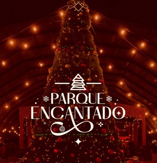 A imagem mostra uma grande árvore de Natal iluminada, decorada com bolas coloridas e luzes douradas. Ela está posicionada no centro de um espaço coberto, com várias luzes penduradas no teto em formato de fios, criando um clima aconchegante e festivo. No centro da imagem, sobre a árvore, há um logotipo branco com o desenho estilizado de uma árvore de Natal e o texto “Parque Encantado” em letras elegantes. O fundo é em tons quentes e avermelhados, transmitindo uma sensação natalina e mágica.