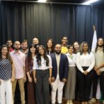 Fotografia de um grupo de cerca de vinte pessoas posando para a câmera em um ambiente interno com cortina preta ao fundo e bandeiras do Brasil e de Pedro Leopoldo à direita. As pessoas estão dispostas em duas fileiras — a da frente em pé e a de trás ligeiramente elevada. A maioria sorri para a foto, vestindo roupas sociais ou casuais elegantes em tons variados de branco, bege, marrom, preto e azul. O cenário sugere um evento institucional, como uma cerimônia de posse ou reunião oficial, transmitindo um clima de união, formalidade e representatividade.