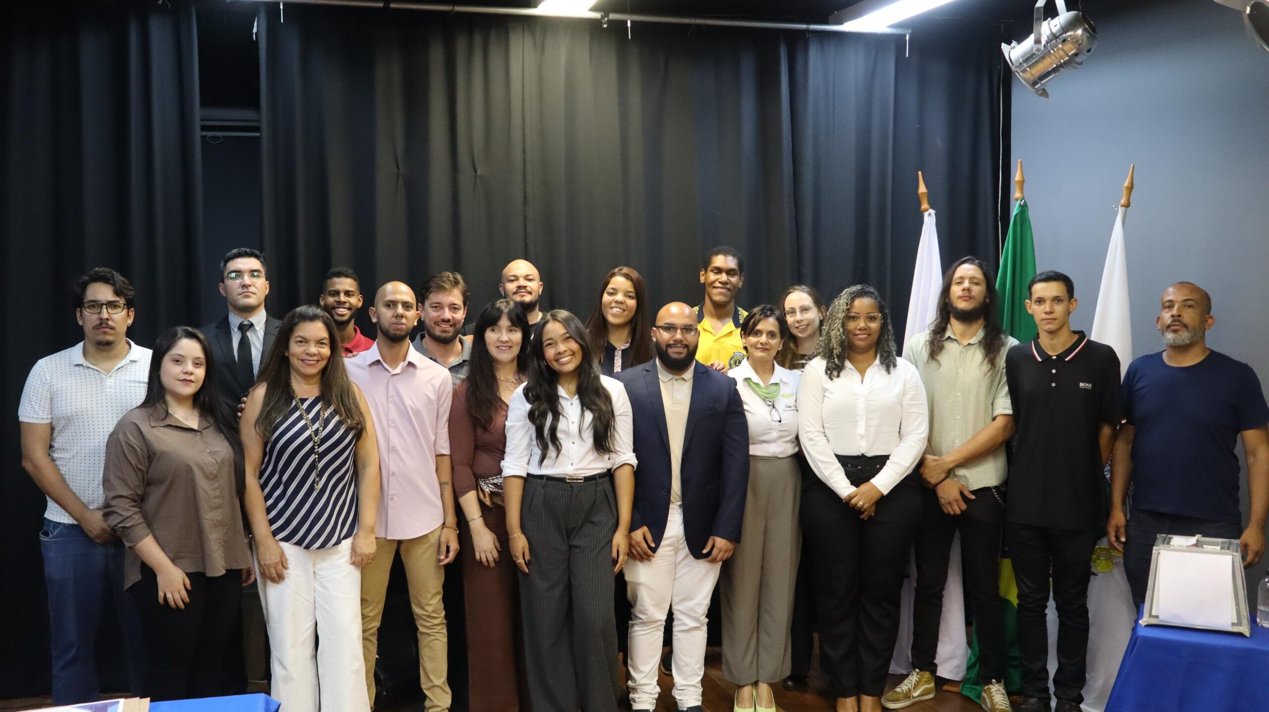 Fotografia de um grupo de cerca de vinte pessoas posando para a câmera em um ambiente interno com cortina preta ao fundo e bandeiras do Brasil e de Pedro Leopoldo à direita. As pessoas estão dispostas em duas fileiras — a da frente em pé e a de trás ligeiramente elevada. A maioria sorri para a foto, vestindo roupas sociais ou casuais elegantes em tons variados de branco, bege, marrom, preto e azul. O cenário sugere um evento institucional, como uma cerimônia de posse ou reunião oficial, transmitindo um clima de união, formalidade e representatividade.